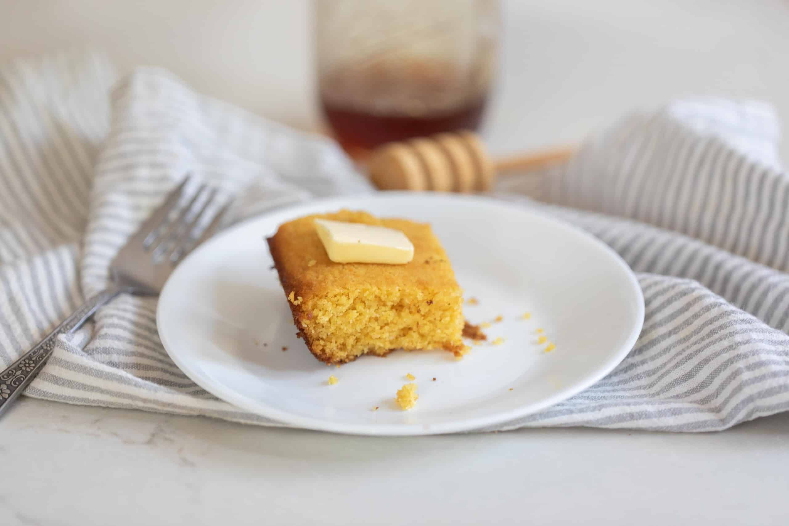a slice of cornbread topped with butter on a white plate on a gray and white stripped towel on a white countertop. A Fork sits to the left and honey is in the background