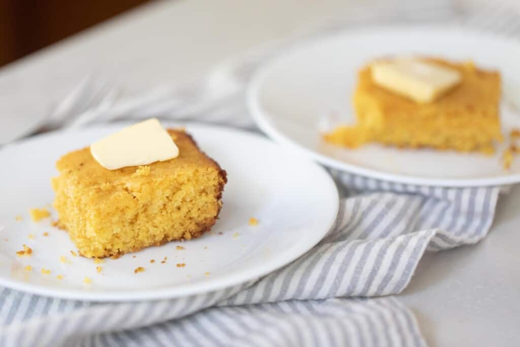 two slices of sourdough cornbread on two separate white plates. A pat of butter tops each slice of bread and the plates sit on a white and gray stripped towel on a white countertop