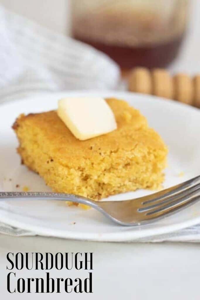 a slice of cornbread topped with butter on a white plate on a gray and white stripped towel on a white countertop. A Fork sits to the left and honey is in the background