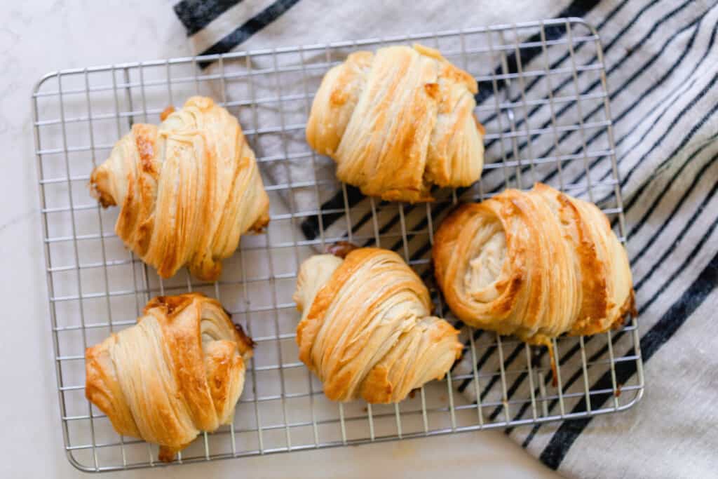 overhead photo of five sourdough croissant rolls on a wire rack that is half on a white and black stripped towel and half on a countertop
