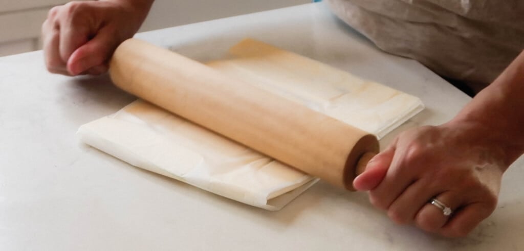 hands holding a rolling pin rolling a butter packet wrapped in parchment paper.