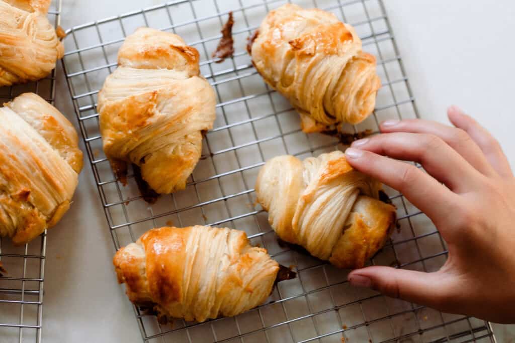 six sourdough crescent rolls on a wire rack. A hand is reaching to pick one up