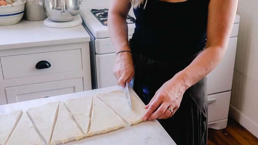 woman wearing a black shirt cutting sourdough croissant dough into triangles with a knife on a white countertop.
