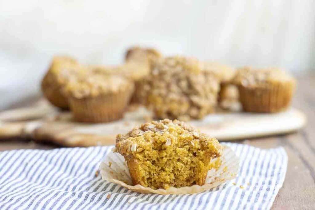 a sourdough pumpkin muffin with a bite take out on a blue and white stripped towel. A cutting board with more muffins ins in the background