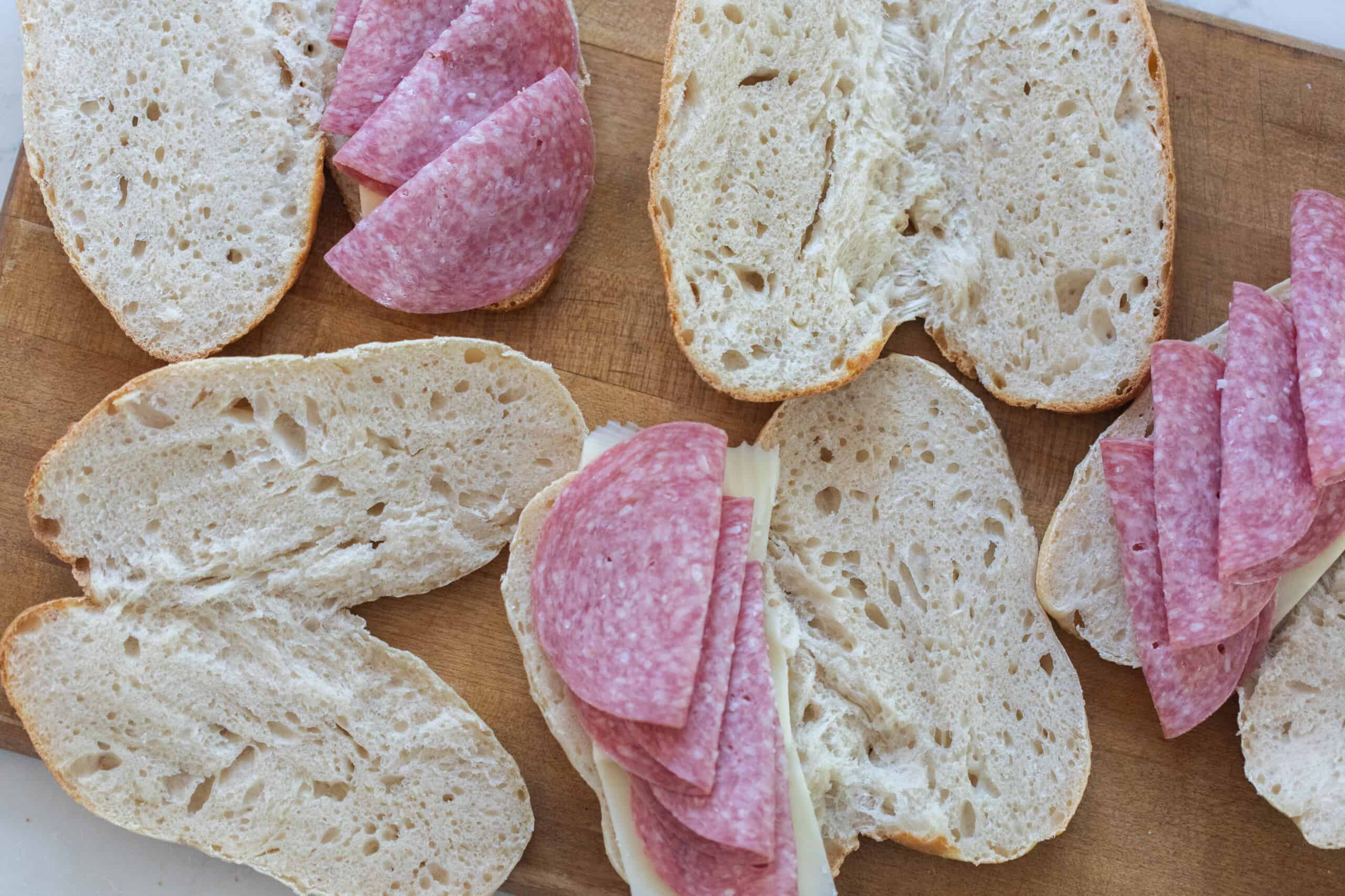 overhead photo of sourdough hoagie rolls sliced in half and topped with cheese and salami on a wood cutting board