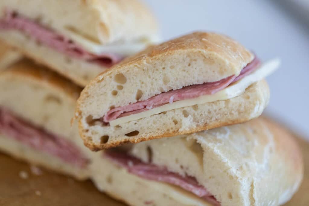 salami and cheese sandwiches on sourdough hoagie rolls sliced in half and stacked on a wood cutting board.