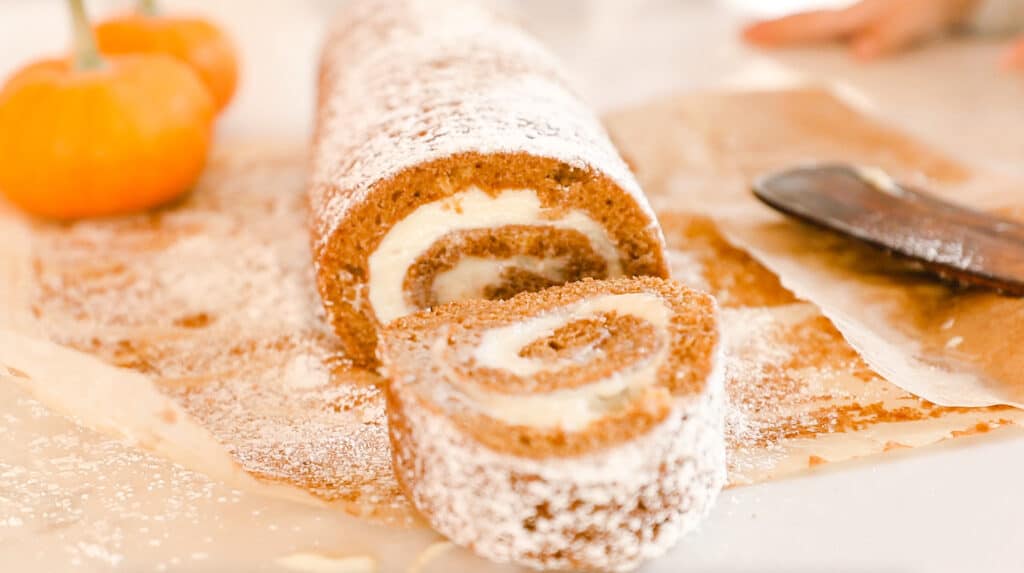 sourdough pumpkin roll with cream cheese frosting filling and dusted with powdered sugar lay on parchment paper with a roll sliced off and resting in front of the roll. A knife is to the right and two small pumpkin to the left