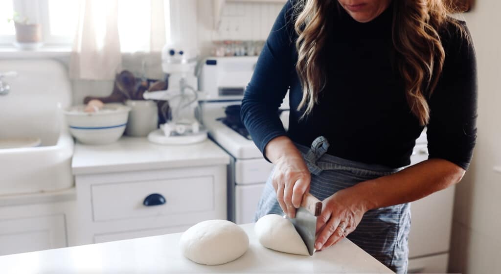 woman wearing a black shirt and blue apron splitting a dough ball in half with a bench scraper on a white counter with a vintage stove and white cabinets in the background