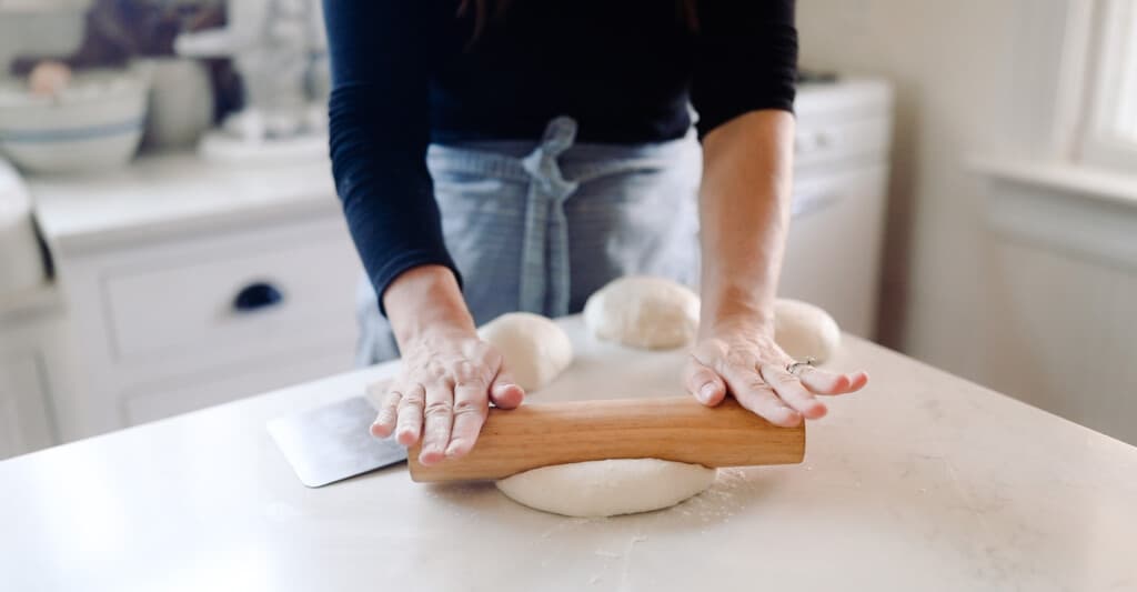 hands pushing a rolling pin into sourdough pizza dough onto a white quartz countertop with more dough balls right in front of the woman.