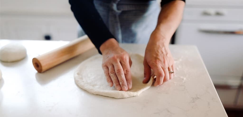 hands pressing out a round sourdough pizza dough making a ridge around the edge on a white quartz countertop