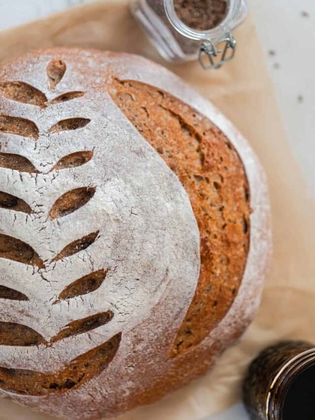 overhead photo of a crusty loaf of sourdough rye bread with a wheat design scored on top. The loaf rest on parchment paper with a jar of spices and a jar of molasses around the loaf