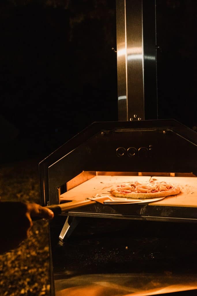 a peel placing a pizza into a outdoor pizza oven on a dark night