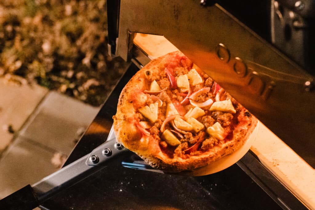 sourdough pizza topped with pineapple, sausage, and onions coming out of a pizza oven with a metal peel