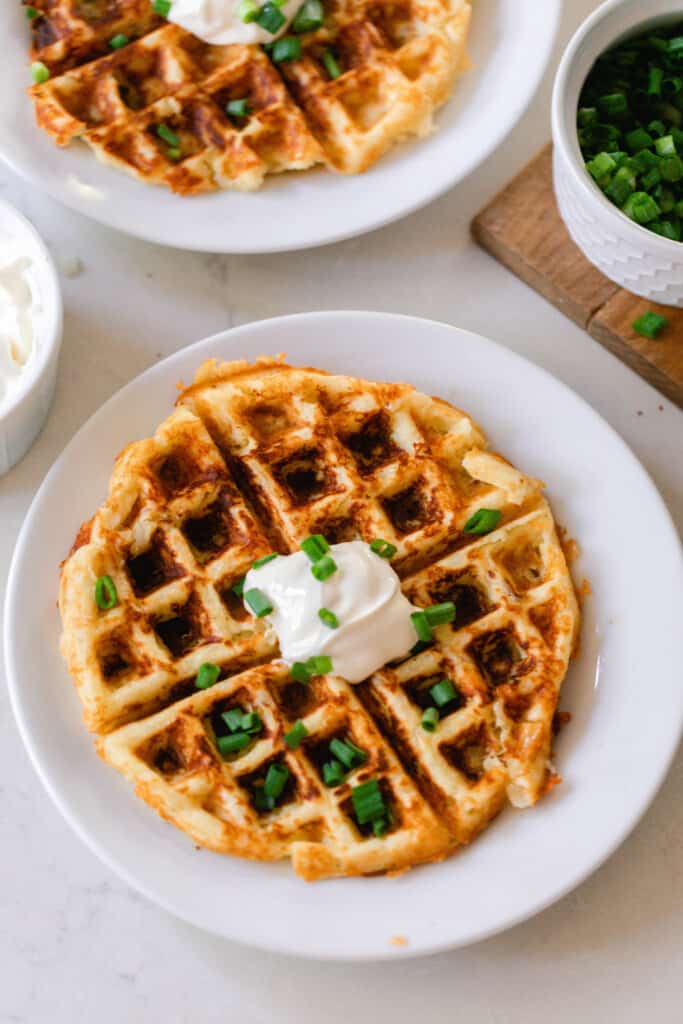 overhead photo of a mashed potato waffle on a white plate topped with sour cream and chives. The plate rests on a white countertop with another potato waffle on a plate in the background