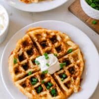 overhead photo of a mashed potato waffle on a white plate topped with sour cream and chives. The plate rests on a white countertop with another potato waffle on a plate in the background