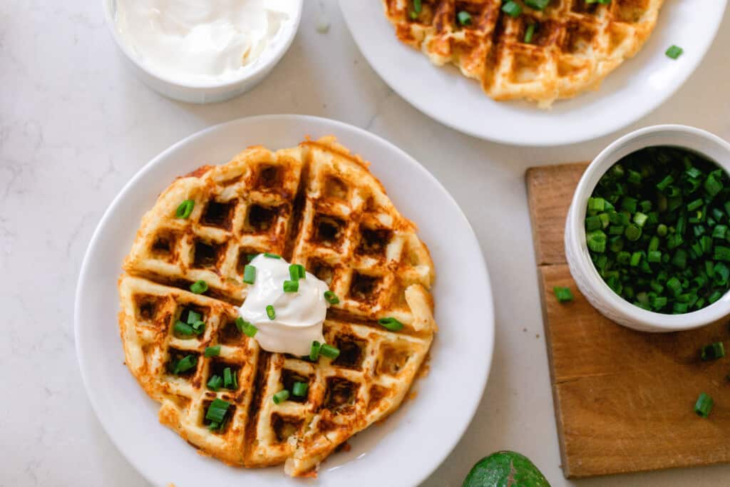 overhead photo two plates of potato waffles on a white countertop with jars of sour cream and chives surrounding the plates.