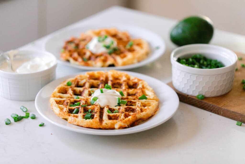 two plates of crispy mashed potato waffles on white plates on a white countertop with jars of toppings around