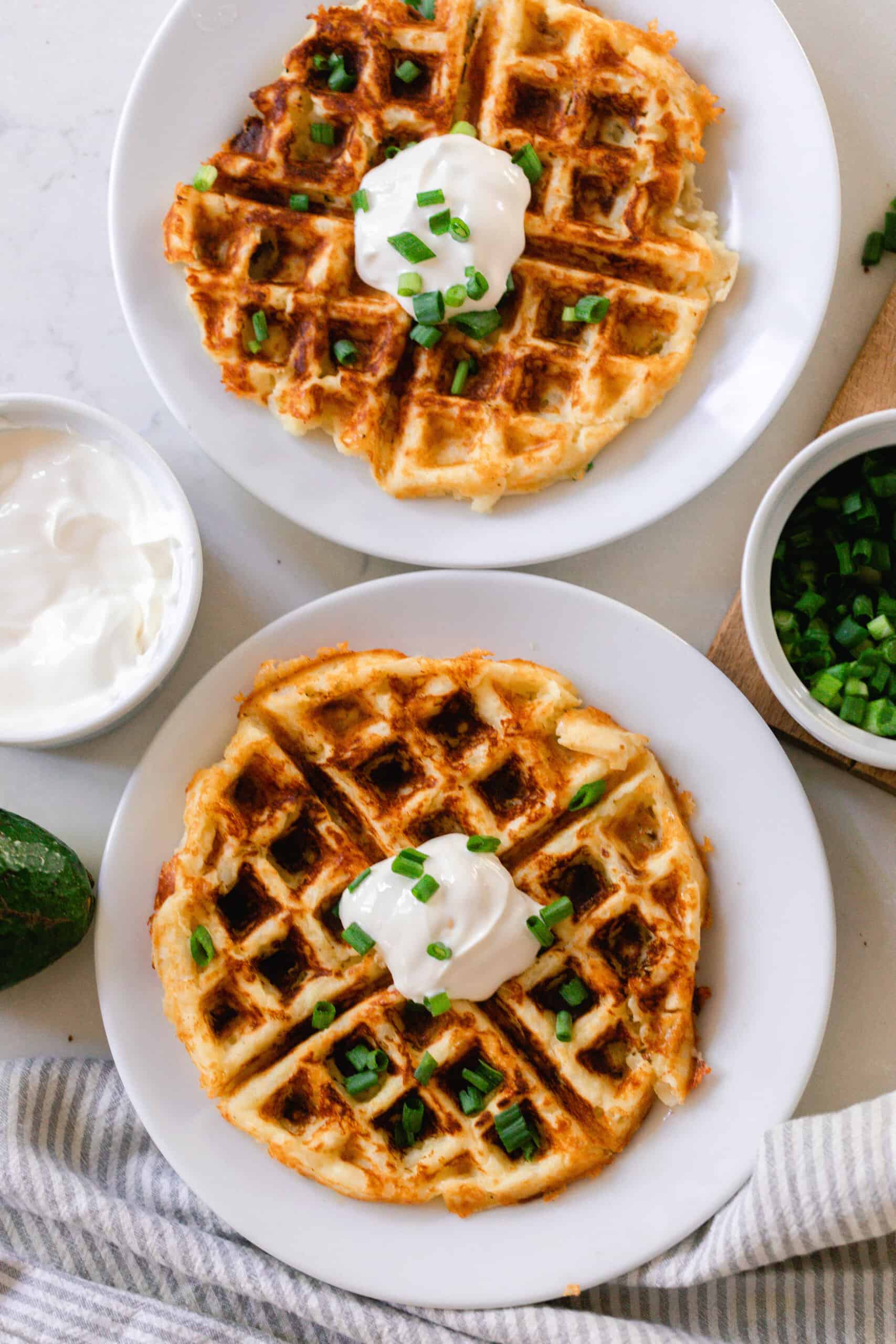 two potato waffles on a white plate topped with sour cream and chives. The plates rest on a white countertop with little jars of chives and sour cream