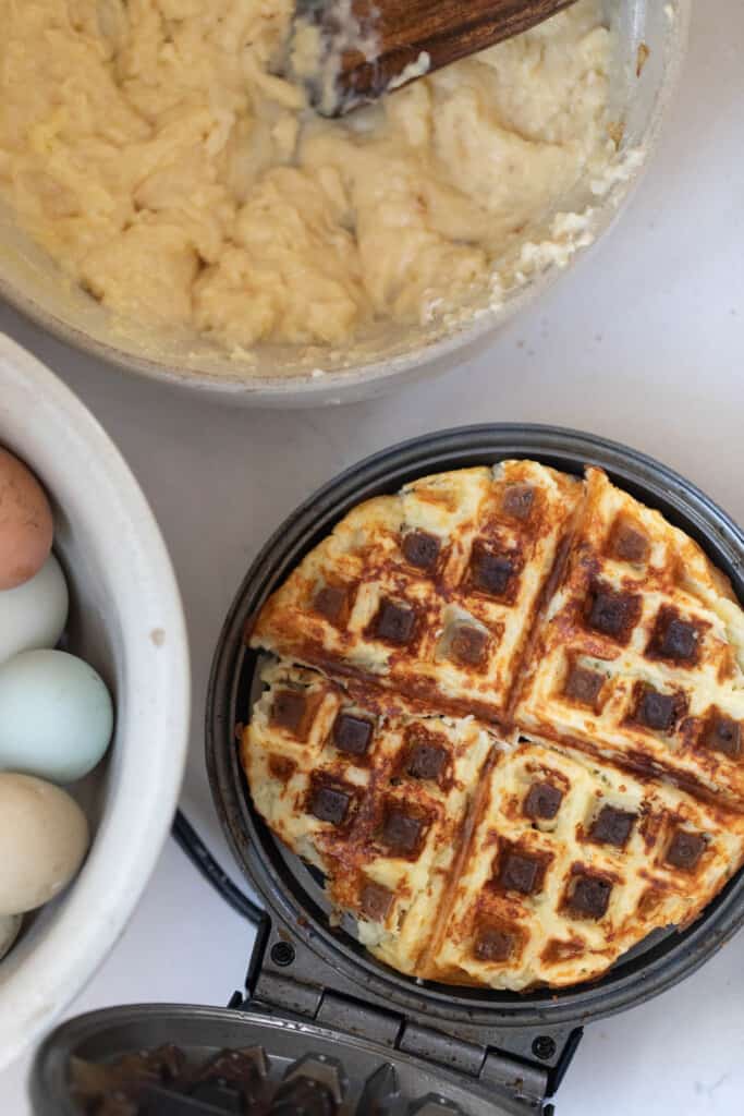 a potato waffle in a waffle maker with a bowl of eggs to the left and a bowl of waffle batter to the top