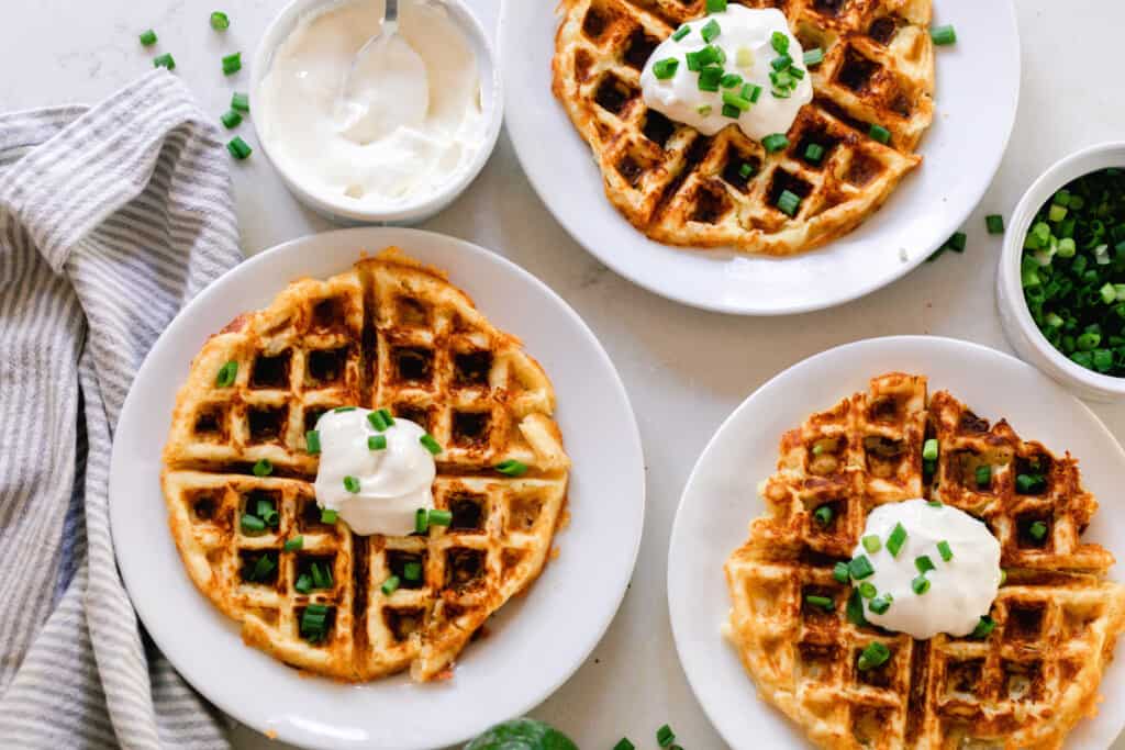 three potato waffles on plates topped with chives and sour cream. With little jars of sour cream and more chives surrounds the plates