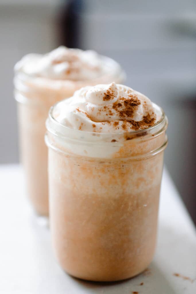 two pumpkin spice smoothies topped with whipped cream and pumpkin spice in glass mason jars on a white countertop