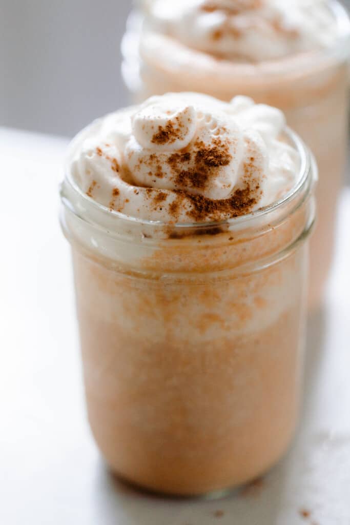 two pumpkin pie smoothies in glass mason jars topped with whipped cream and sprinkled with more pumpkin spice