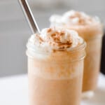 two pumpkin smoothies topped with whipped cream sprinkled with pumpkin spice. A metal straw is in the smoothie and the smoothies sit on a white countertop