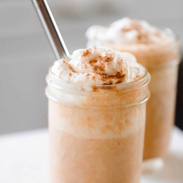 two pumpkin smoothies topped with whipped cream sprinkled with pumpkin spice. A metal straw is in the smoothie and the smoothies sit on a white countertop