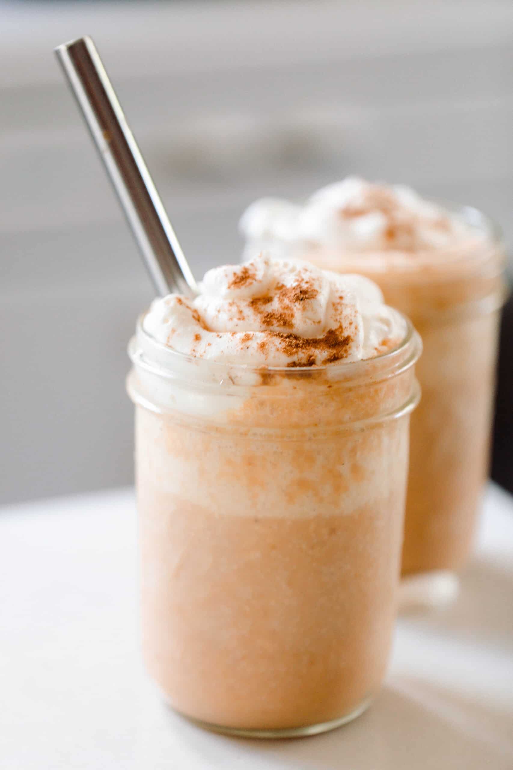 two pumpkin smoothies topped with whipped cream sprinkled with pumpkin spice. A metal straw is in the smoothie and the smoothies sit on a white countertop