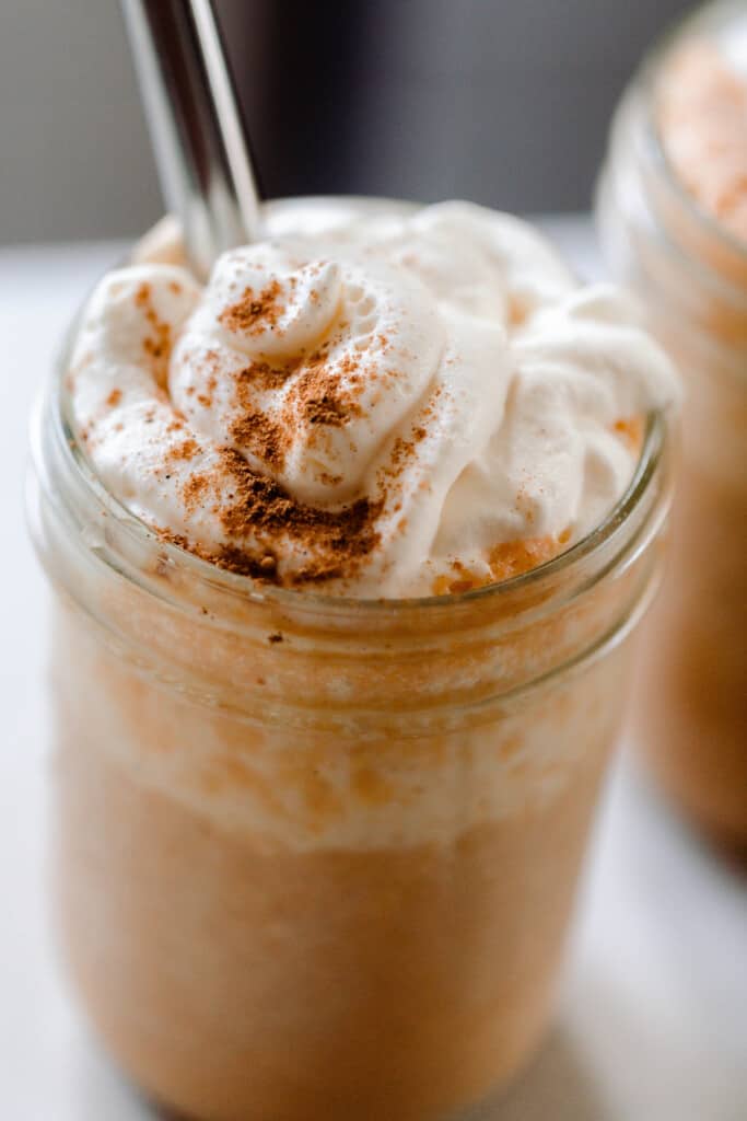 close up picture of whipped cream sprinkled with pumpkin spice on top of a pumpkin smoothie in a glass mason jar with a metal straw.