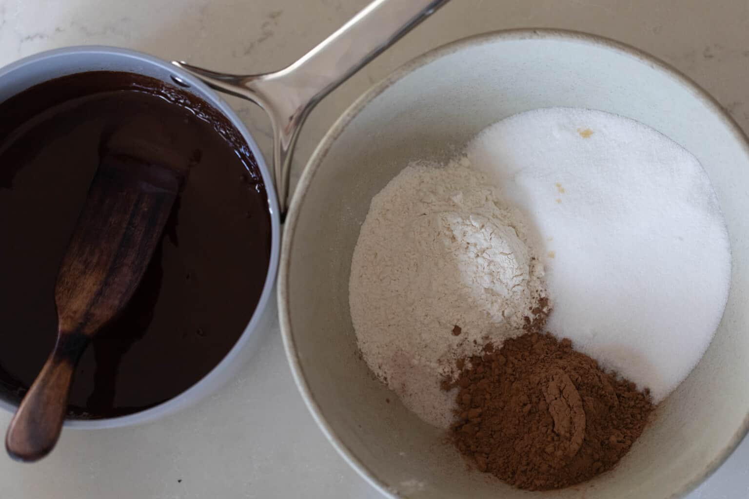 Cocoa powder, sugar, and flour in a bowl. A pot of melted chocolate is to the left.