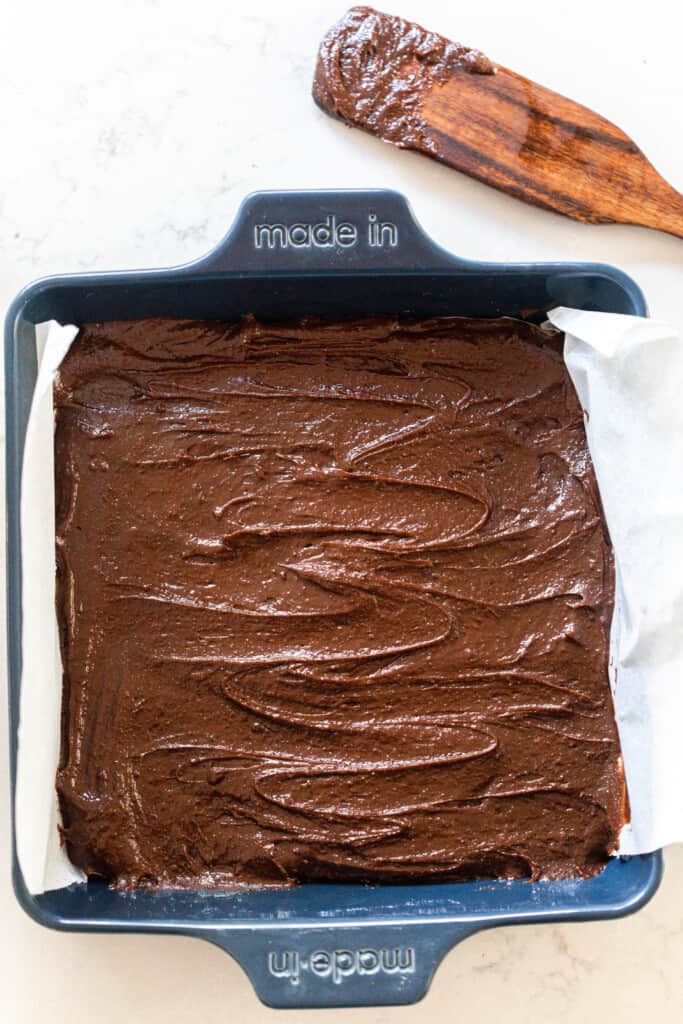 overhead photo of sourdough brownie batter poured into a blue 9x0 baking dish with a wooden spatula in the back right corner