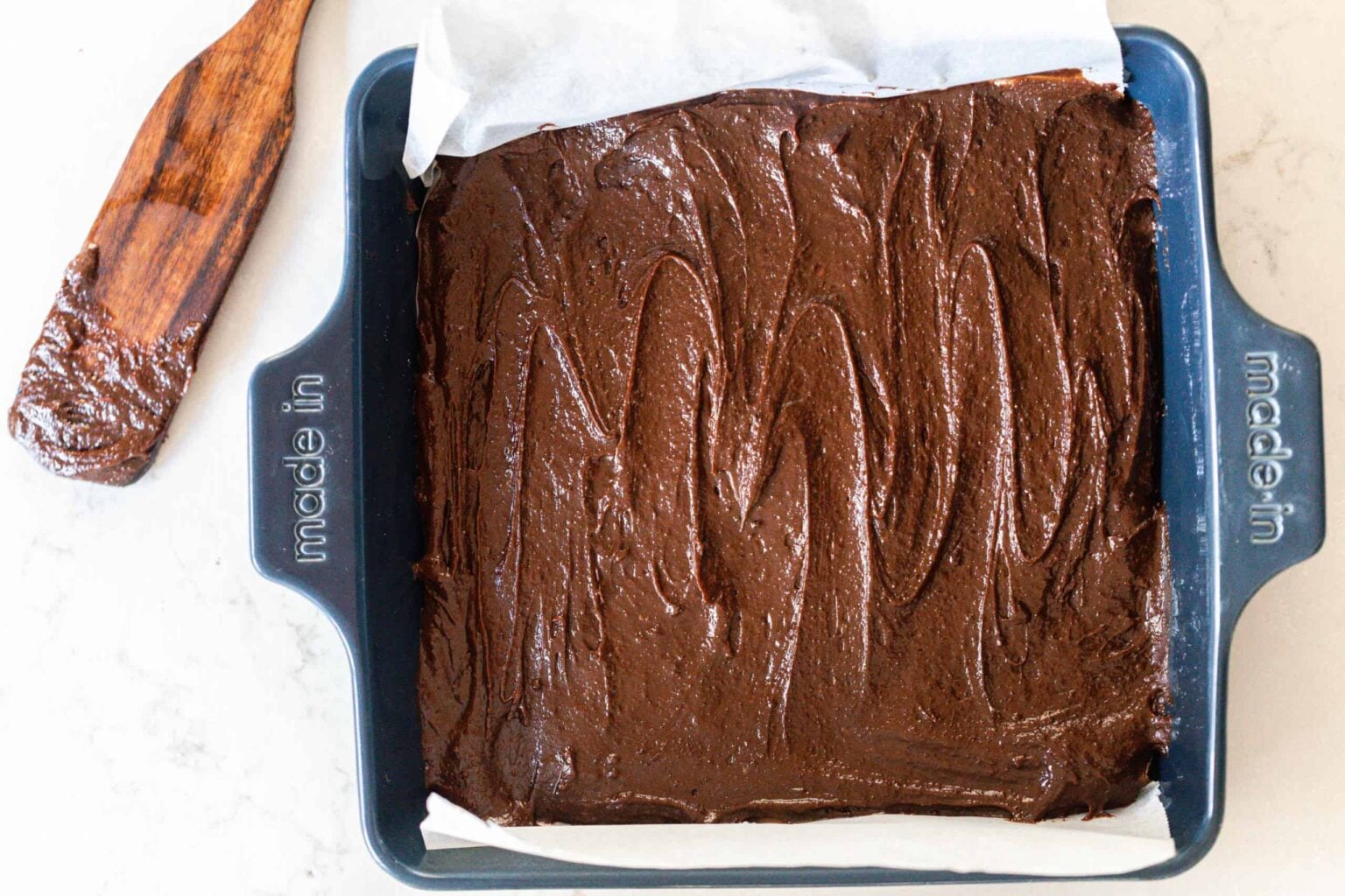 Overhead photo of sourdough brownie batter poured into a blue 9x9 baking dish with a wooden spatula in the back right corner.