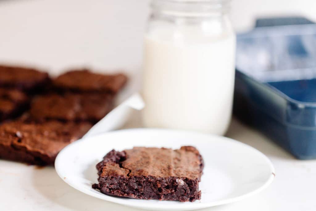 a square of brownie on a white plate with a glass of milk and the remaining brownies behind the plate.