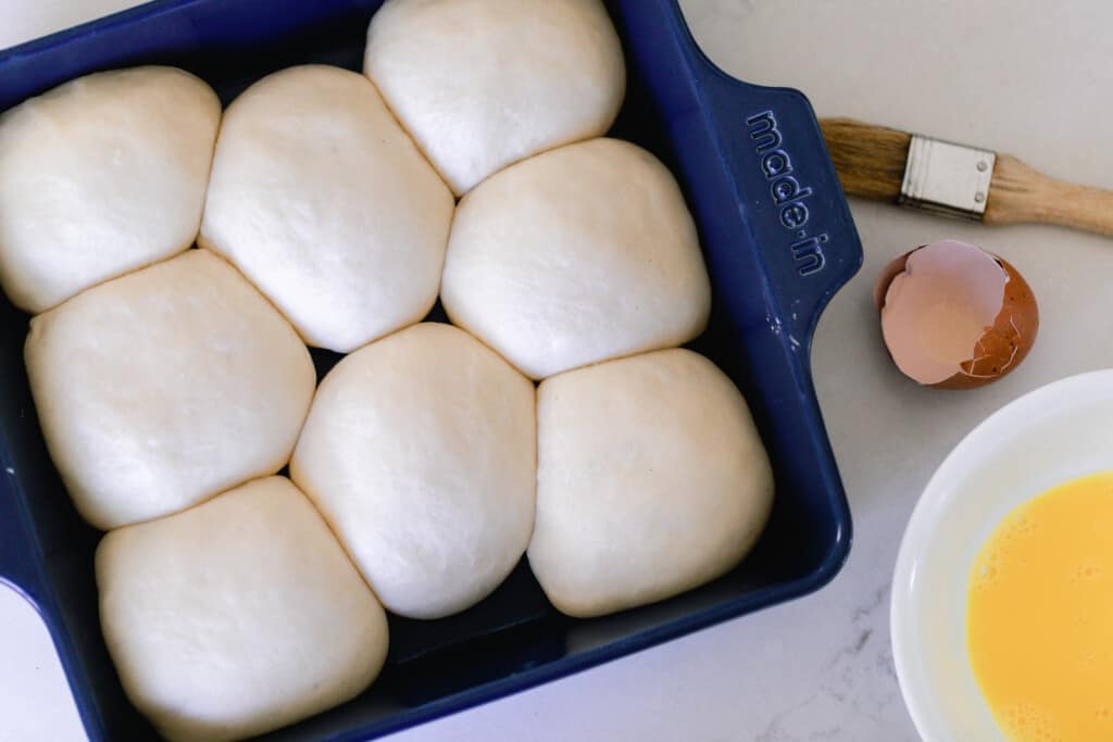 overhead photo of 8 sourdough dinner rolls in a navy 8x8 baking dish with a pastry brush and a small bowl of melted butter to the right