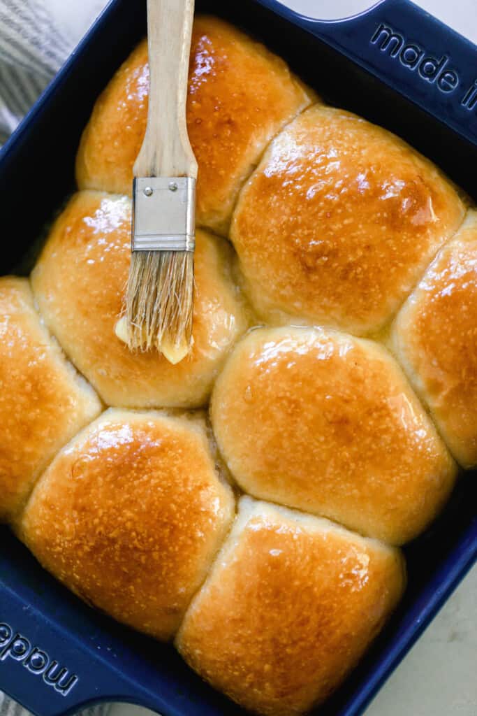 overhead photo of sourdough dinner rolls baked in a navy baking dish with a dark golden color. A pastry brush with melted butter is on top