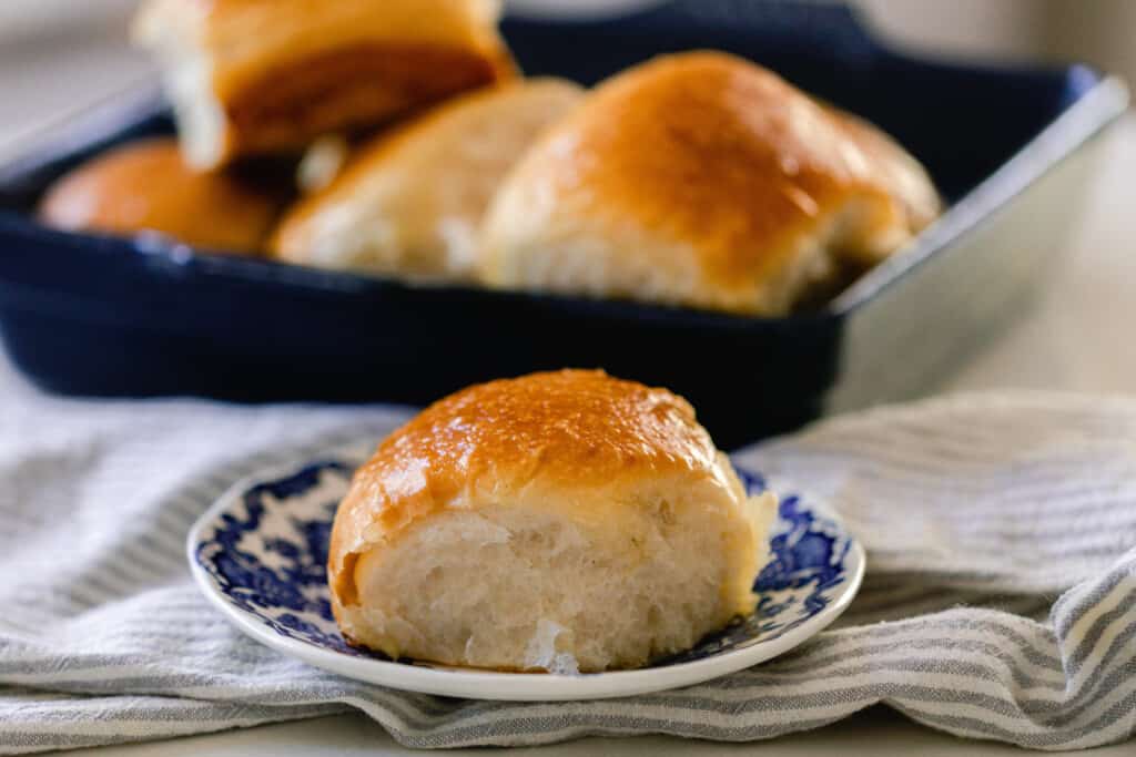 sourdough dinner roll with a golden crust sits on a vintage blue and white plate on a cream towel with a navy baking dish full of rolls in the background