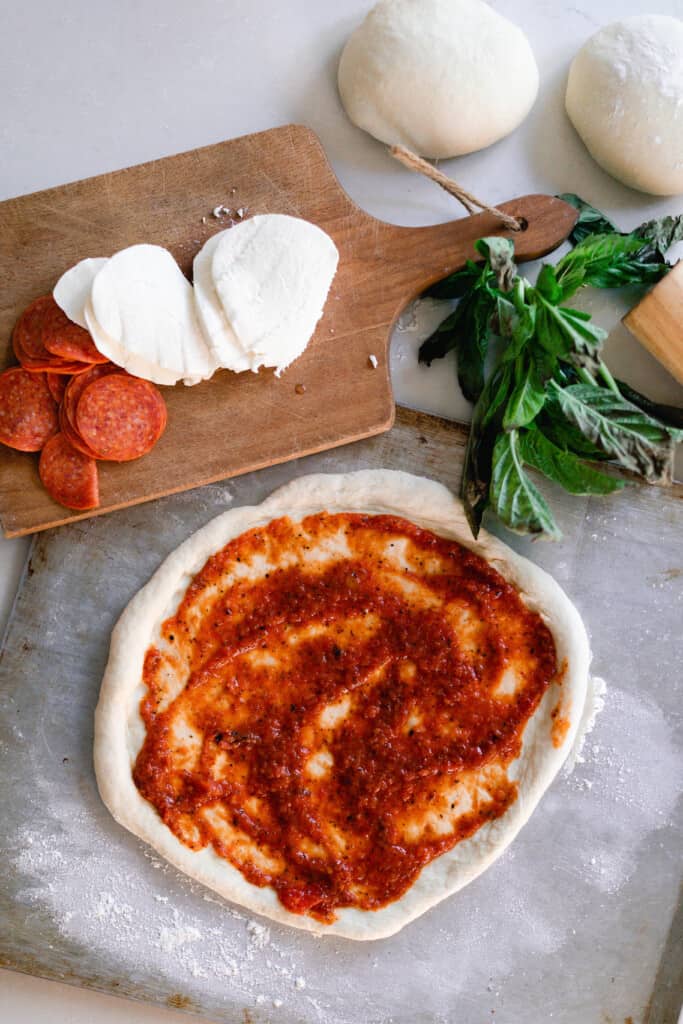 sourdough pizza dough rolled out on a flat cookie sheet with pizza sauce spread over top. A cutting board in the back has fresh mozzarella and pepperonis. More basil and pizza dough balls to the right of the cutting board.