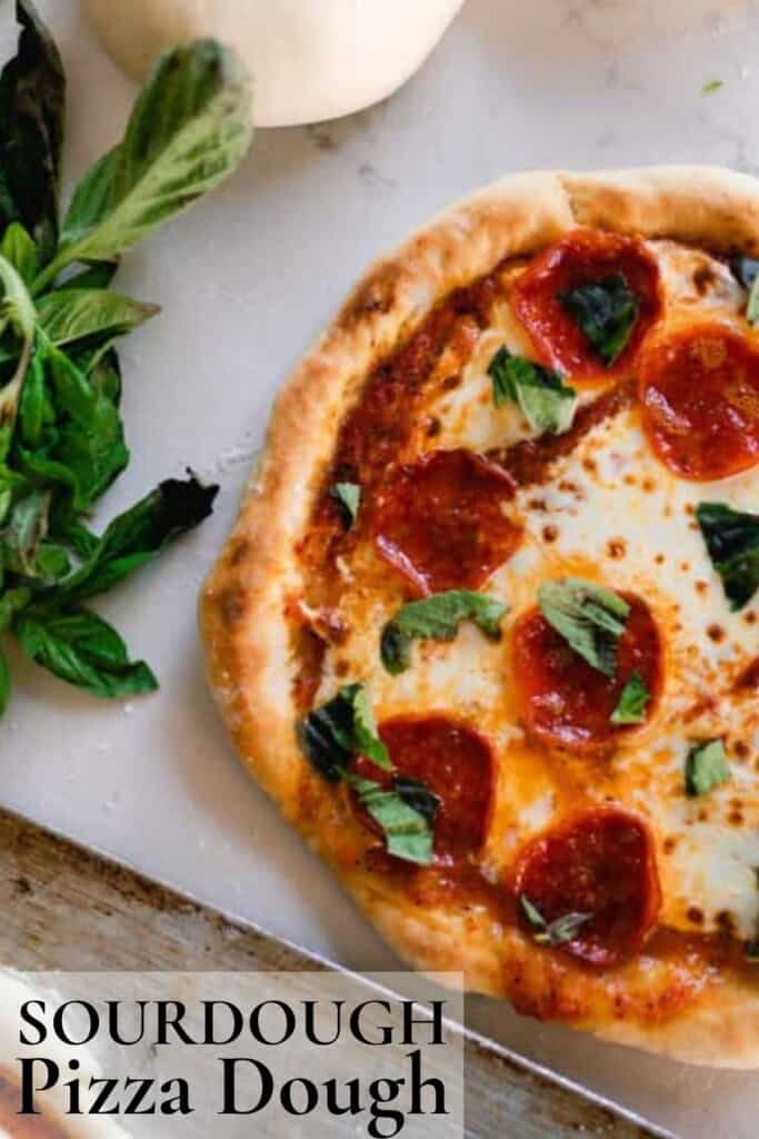 half of a sourdough pizza topped with sauce, cheese, pepperonis, and basil on a white countertop with fresh basil to the left