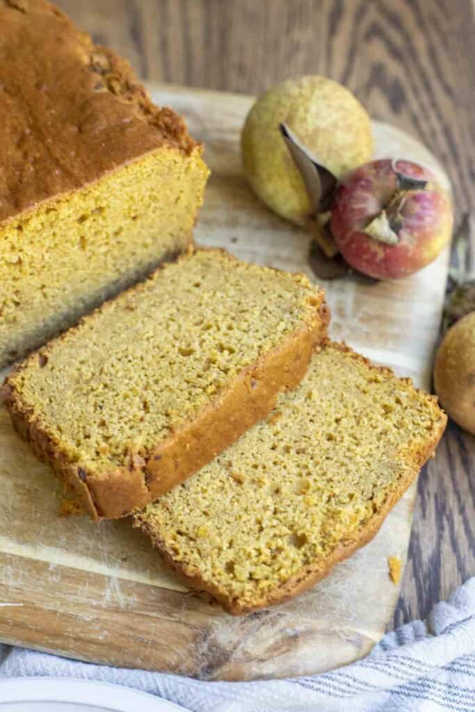 sourdough pumpkin bread with two slices laying on a wood cutting board with an apple and bear in the back right corner