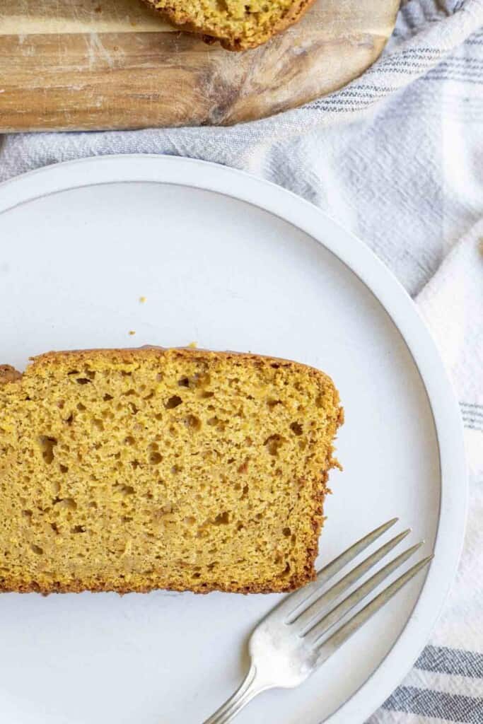 close up picture of a slice of sourdough pumpkin bread on a cream colored plate on a white and blue stripped towel with a wood cutting board in the background