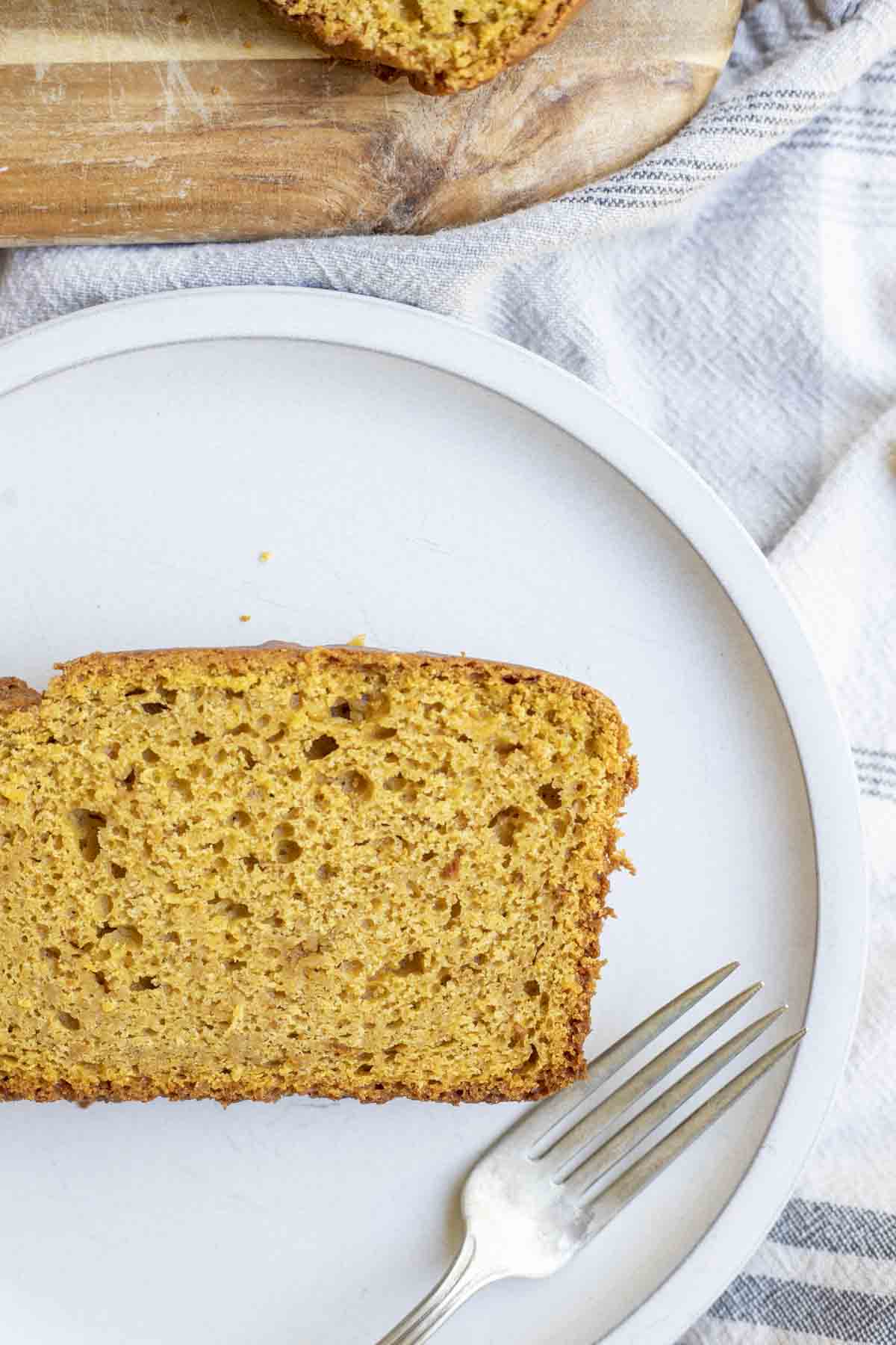 close up picture of a slice of sourdough pumpkin bread on a cream colored plate on a white and blue stripped towel with a wood cutting board in the background