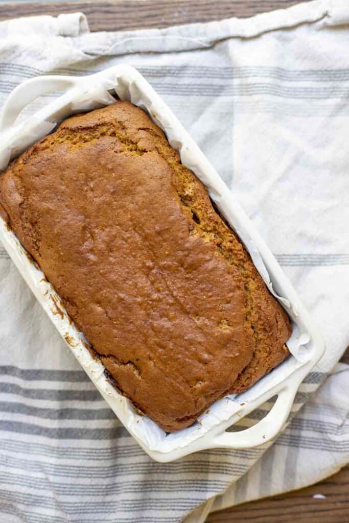 overhead photo of a loaf of sourdough pumpkin bread in a parchment lined stone bread pan on top of a blue and white stripped towel on a wood table