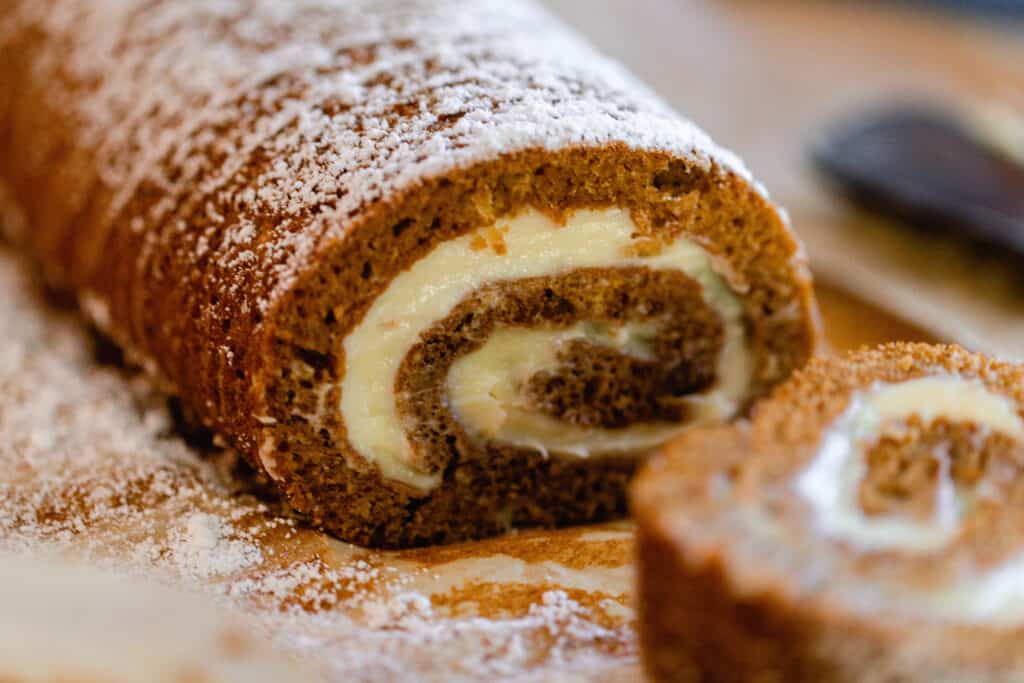 sourdough pumpkin roll with cream cheese frosting filling dusted with powdered sugar on parchment paper with a slice of the roll to the front right