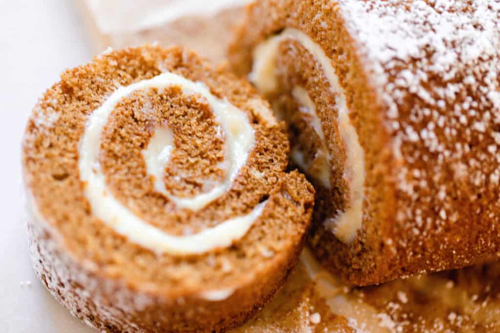 a slice of a sourdough pumpkin roll with cream cheese filling rolled up in pumpkin cake. The rest of the roll is to the right and dusted with powdered sugar.