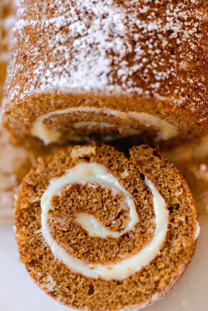 overhead photo of a sourdough pumpkin roll dusted with powdered sugar and one slice revealing the roll and cream cheese frosting filling on a white countertop