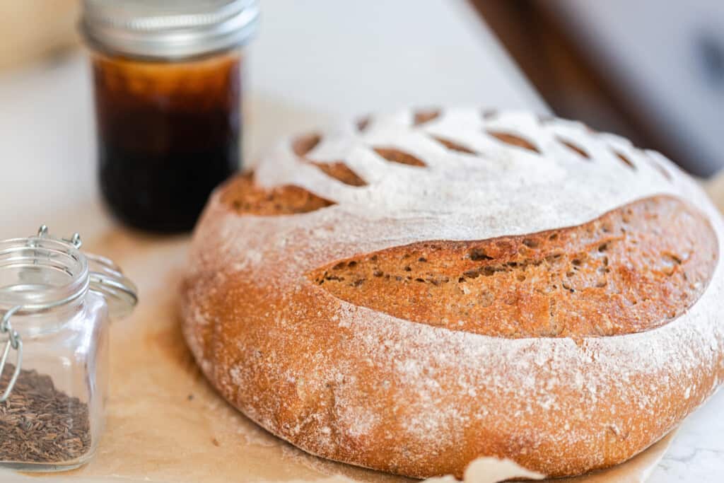 loaf of sourdough rye bread with the top dusted with flour with a wheat pattern on top. The loaf sits on the cutting board with a jar of molasses and a little glass of seasonings beside it