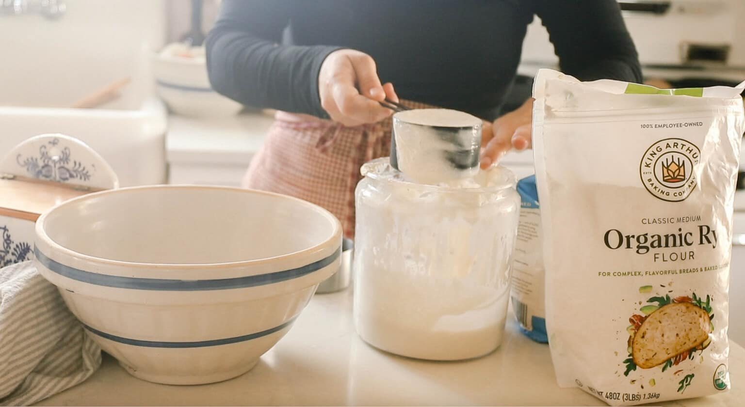 woman wearing a black shirt and red half apron removing sourdough starter from a large jar. A large cream bowl with blue stripes is to the left and a bag of flour to the right.