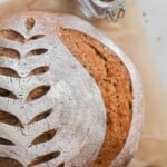 overhead photo of a crusty loaf of sourdough rye bread with a wheat design scored on top. The loaf rest on parchment paper with a jar of spices and a jar of molasses around the loaf