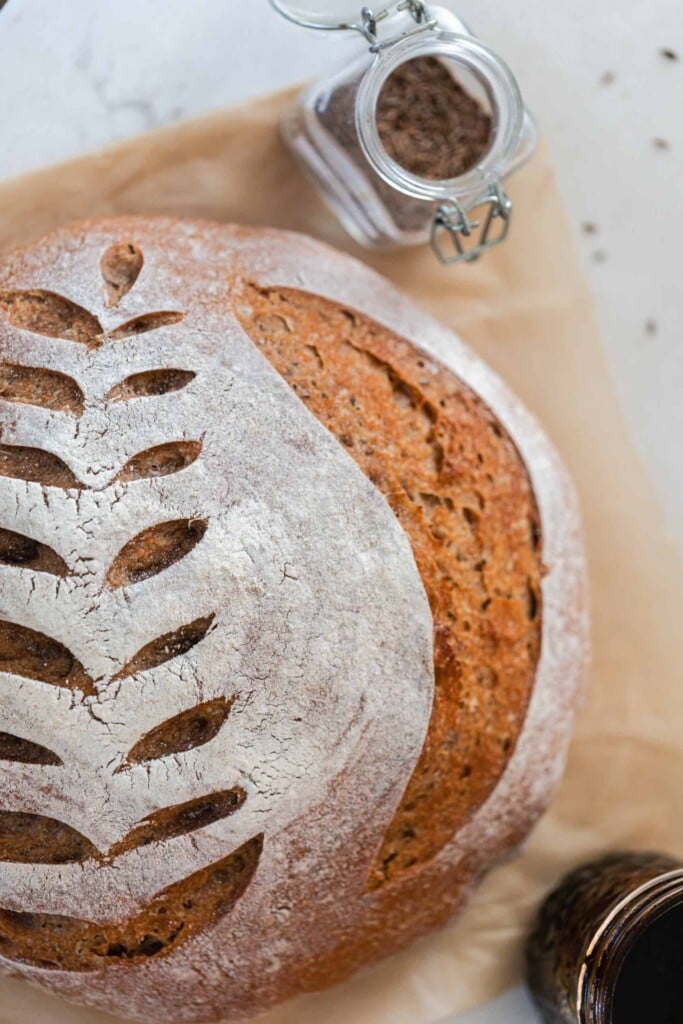 overhead photo of a crusty loaf of sourdough rye bread with a wheat design scored on top. The loaf rest on parchment paper with a jar of spices and a jar of molasses around the loaf